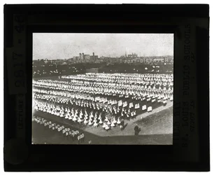 Horizontales, schwarz-weißes Foto von Hunderten von Schulkindern, die für die Feldtagparade und das Fest in Formation stehen. Alle Mädchen in weißen Kleidern und die Jungen in weißen Hemden und Krawatten mit dunklen Hosen